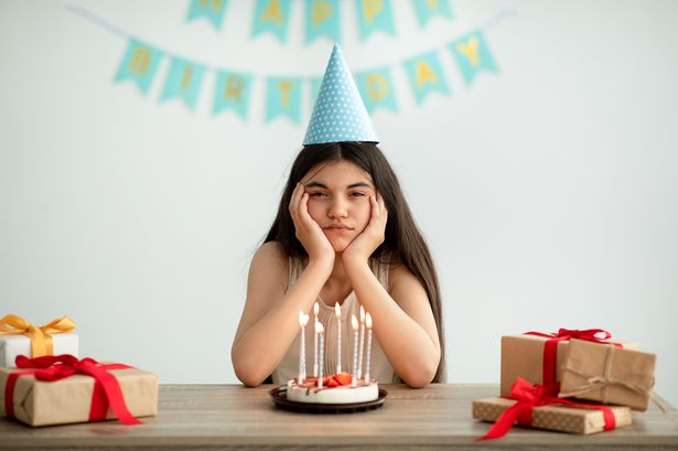 0 Portrait of disappointed Indian teen girl in party hat sitting alone at table with gifts and birthda